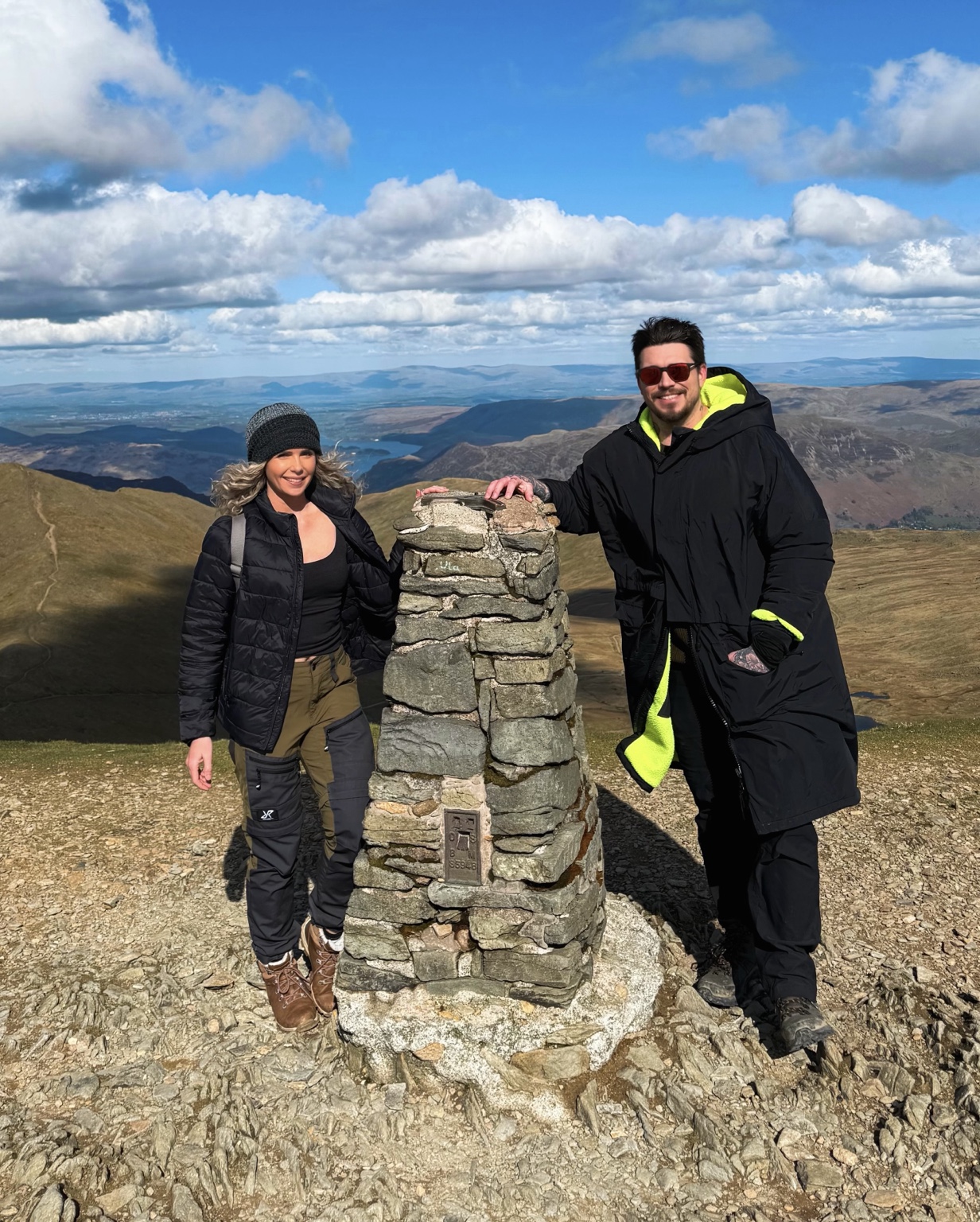 Lana and Tomm standing together beside a mountain summit marker
